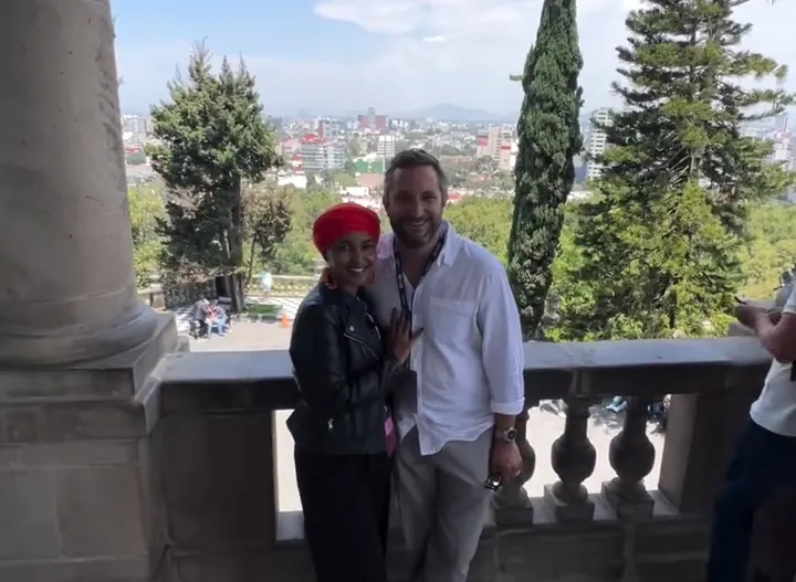 Ilhan Omar and Tim Mynett smiling together with a city skyline in the background.