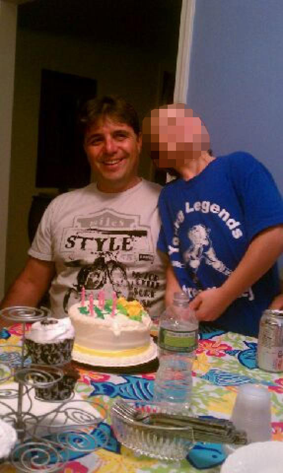 A smiling man and a child with a blurred face stand behind a birthday cake with lit candles.