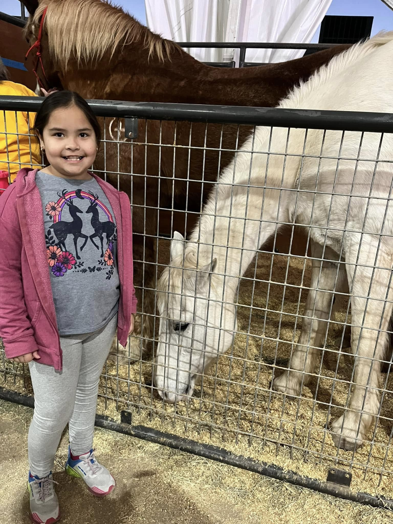 A young girl smiling in front of a white horse in an enclosure, with a brown horse visible in the background.