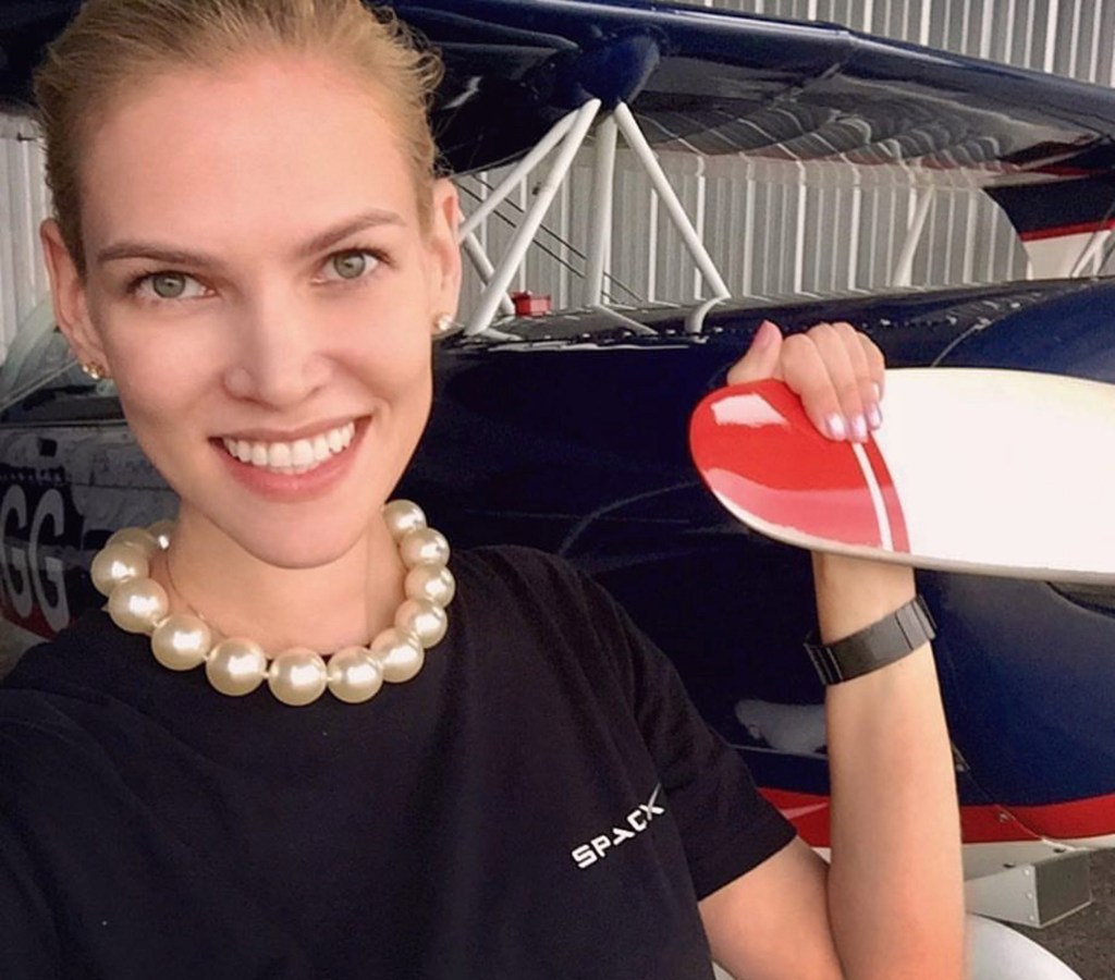 Nadia Marcinko, pilot, wearing a pearl necklace and a black t-shirt, stands in front of a blue airplane and holds part of the plane's propeller.