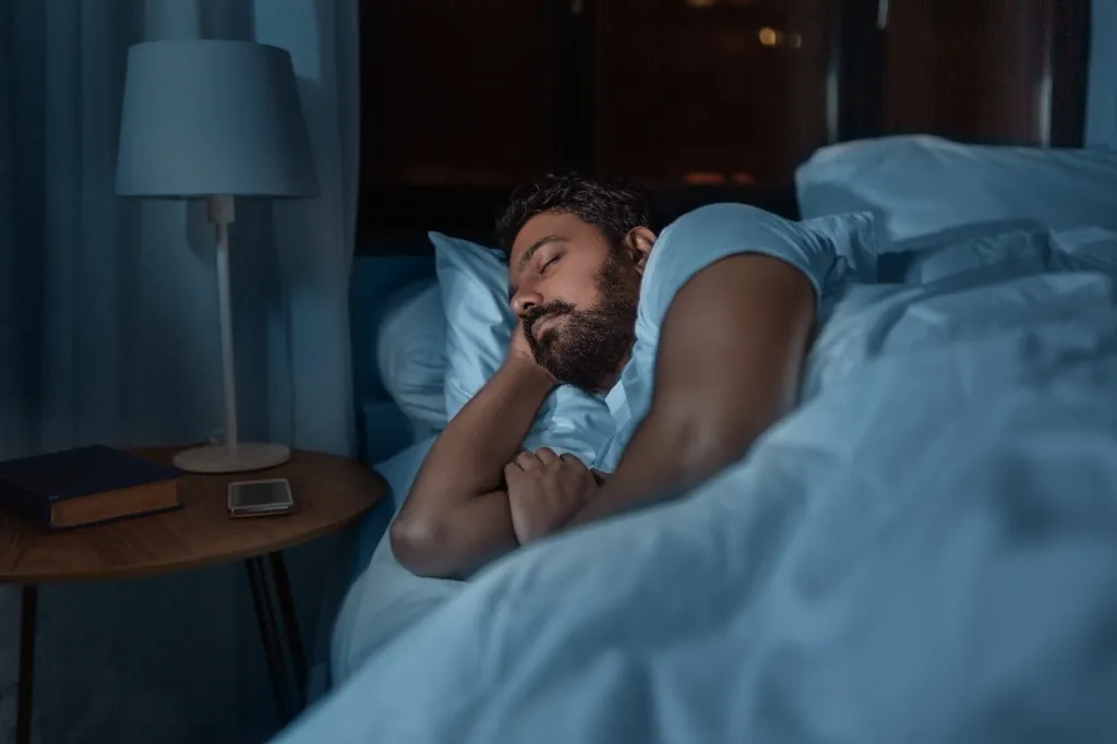 A man with a beard is asleep in bed at night, with a lamp, book, and phone on a bedside table.