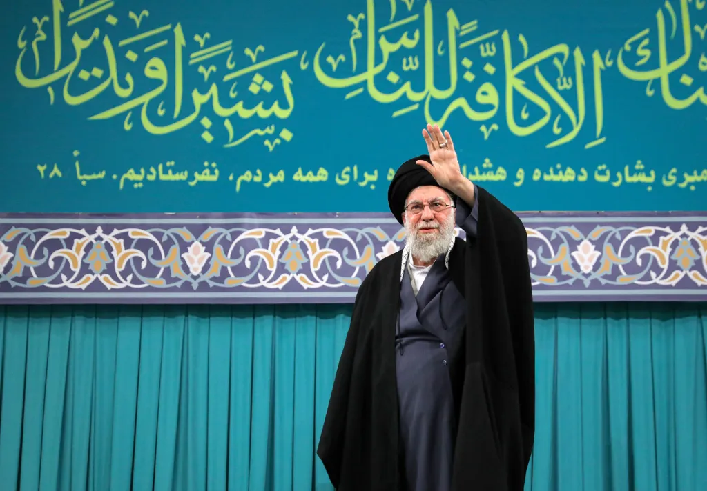 The Iranian Supreme Leader raises his hand wearing all black against a green wall. 