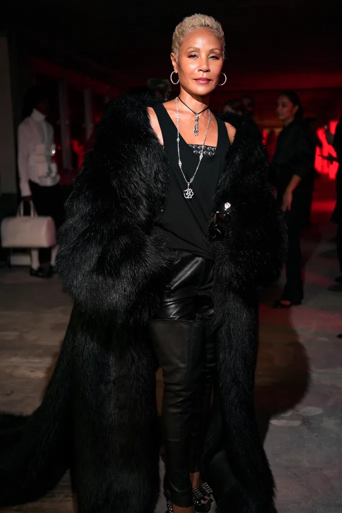 Jada Pinkett Smith in a black fur coat, black top, and black leather pants, with multiple necklaces and hoop earrings, posing for a photo.