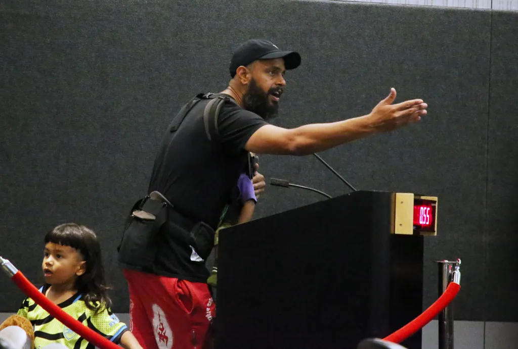 A man speaking at a podium with a young child nearby at a Police Commission meeting.