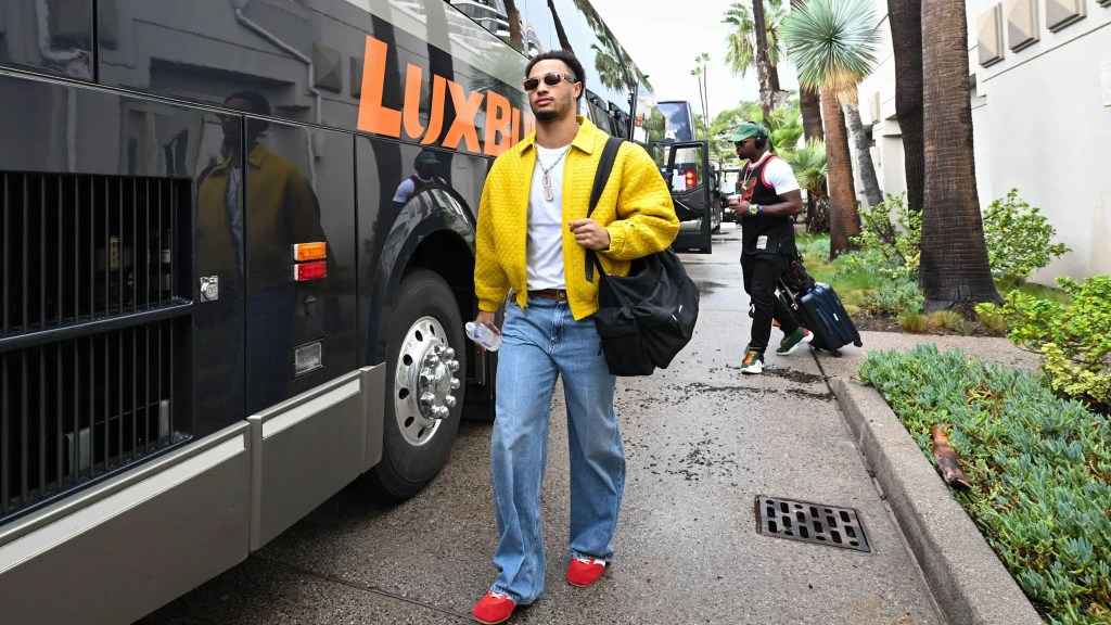 Jaxon Smith-Njigba wearing a yellow jacket, light blue jeans, and red shoes, walking past a black bus.