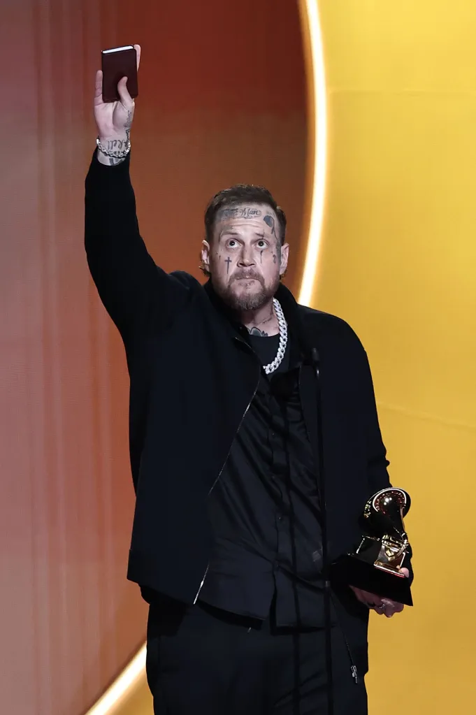 Jelly Roll holding a Grammy award and a small red item in his raised hands.
