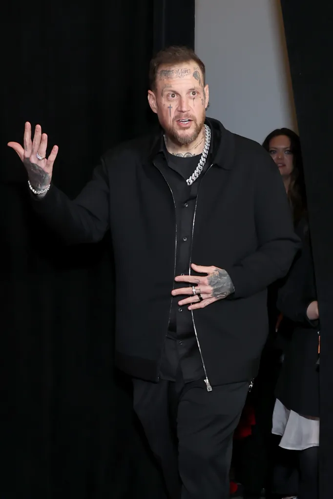 Jelly Roll, winner of multiple GRAMMY Awards, wearing a black shirt and jacket, waving to the camera.