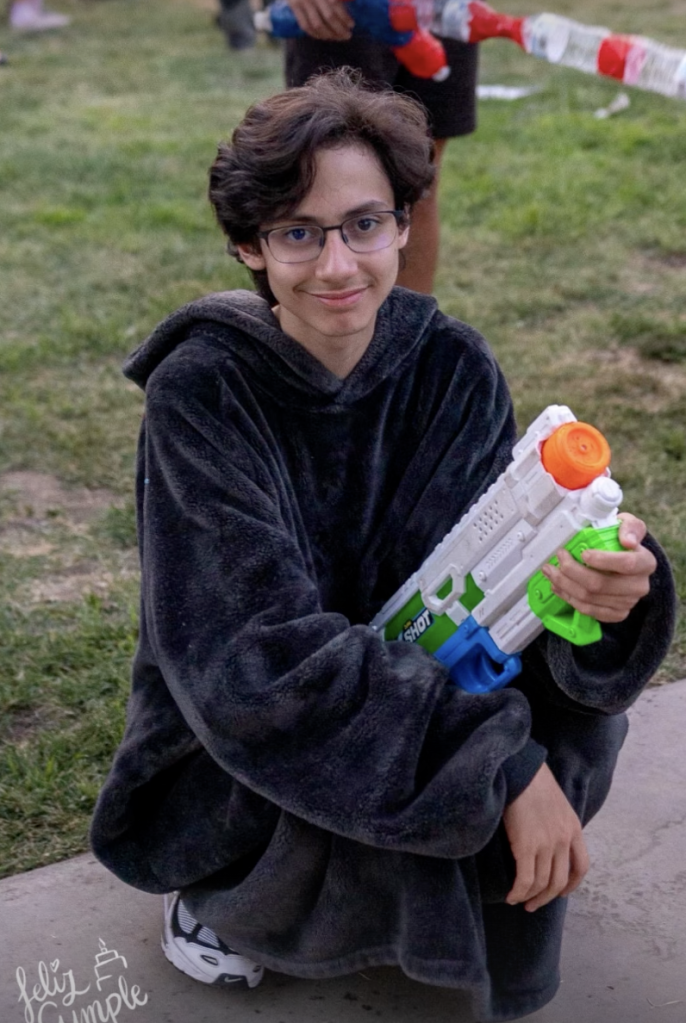 Jennifer Lopez's child, Max, squats outdoors holding a toy water gun and smiling at the camera.
