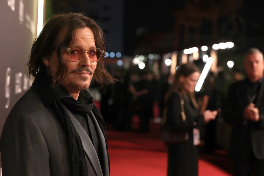 Johnny Depp with shoulder-length hair and a mustache smiles at the camera, wearing pink-tinted glasses, a black suit, and a dark scarf.