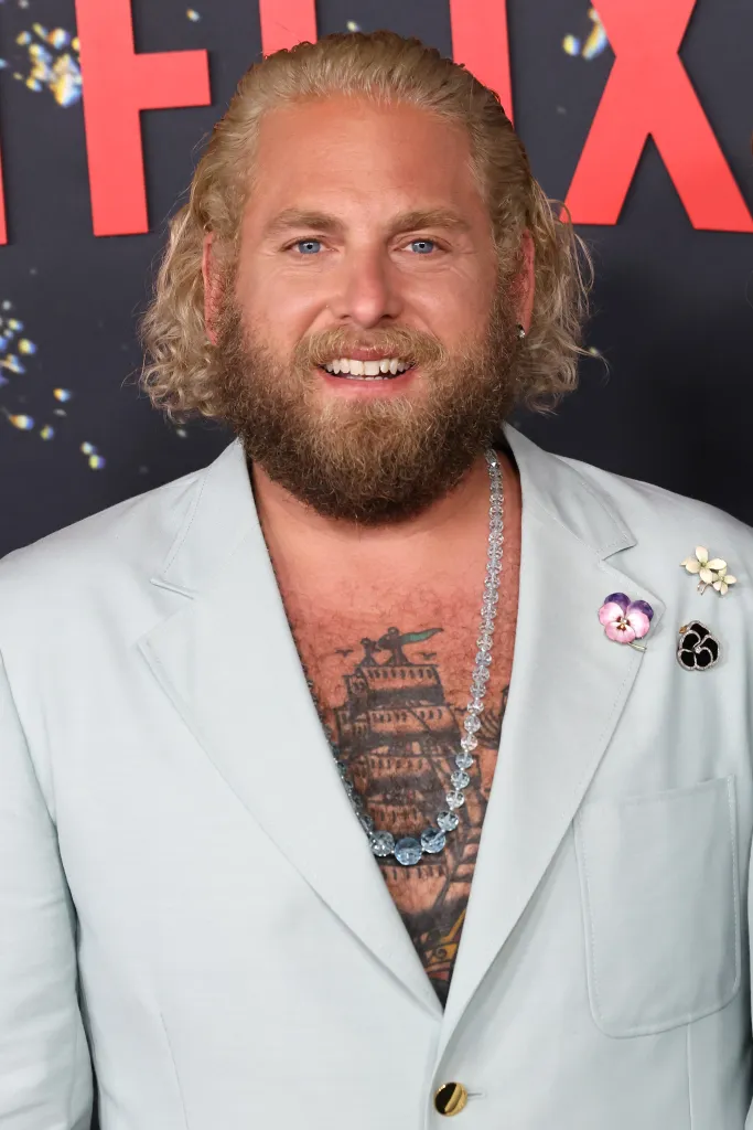 Jonah Hill smiling, wearing a light blue suit jacket open to reveal a tattooed chest and a beaded necklace, with long blond hair and beard.