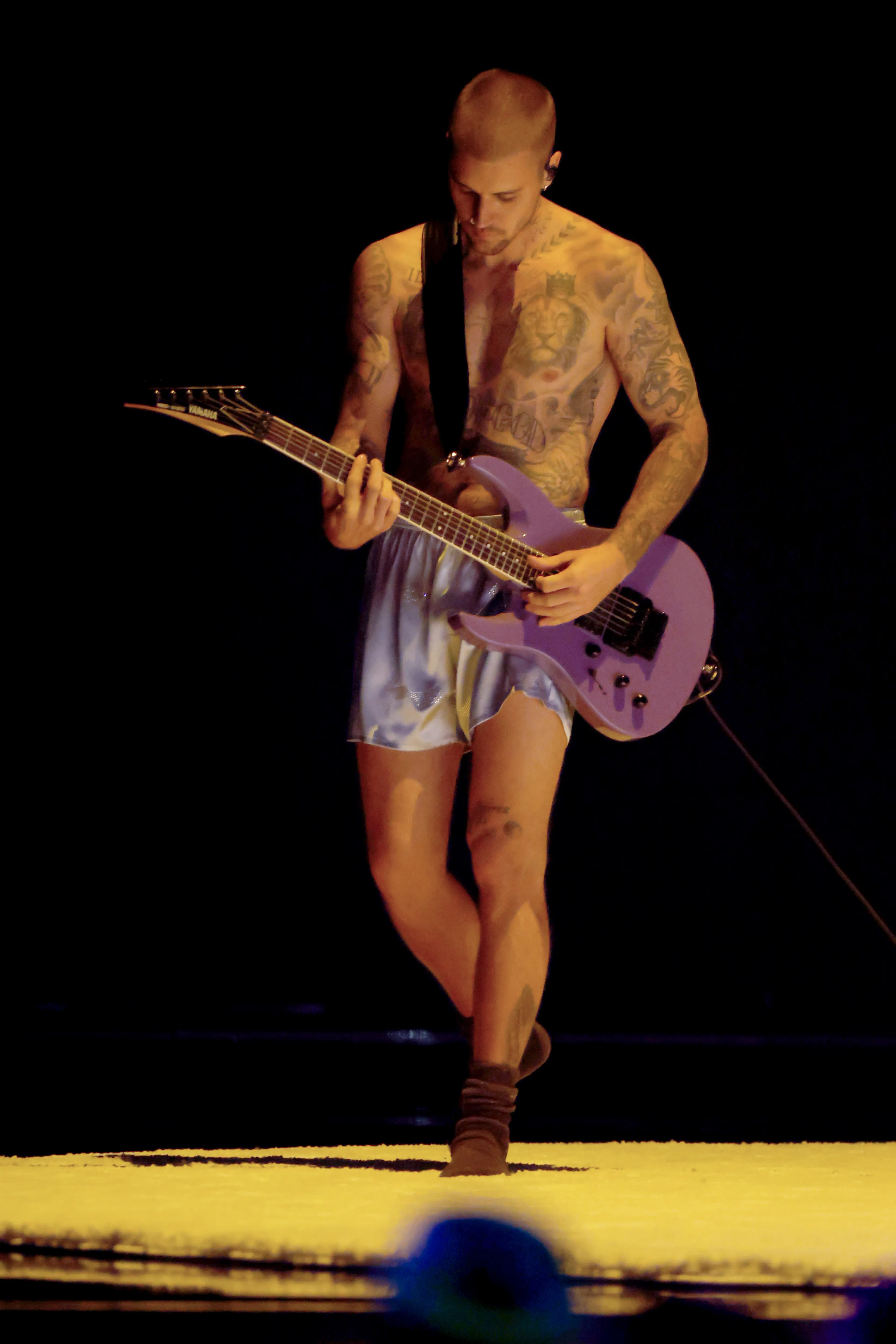 Justin Bieber performing on stage, shirtless with a purple guitar and satin shorts.
