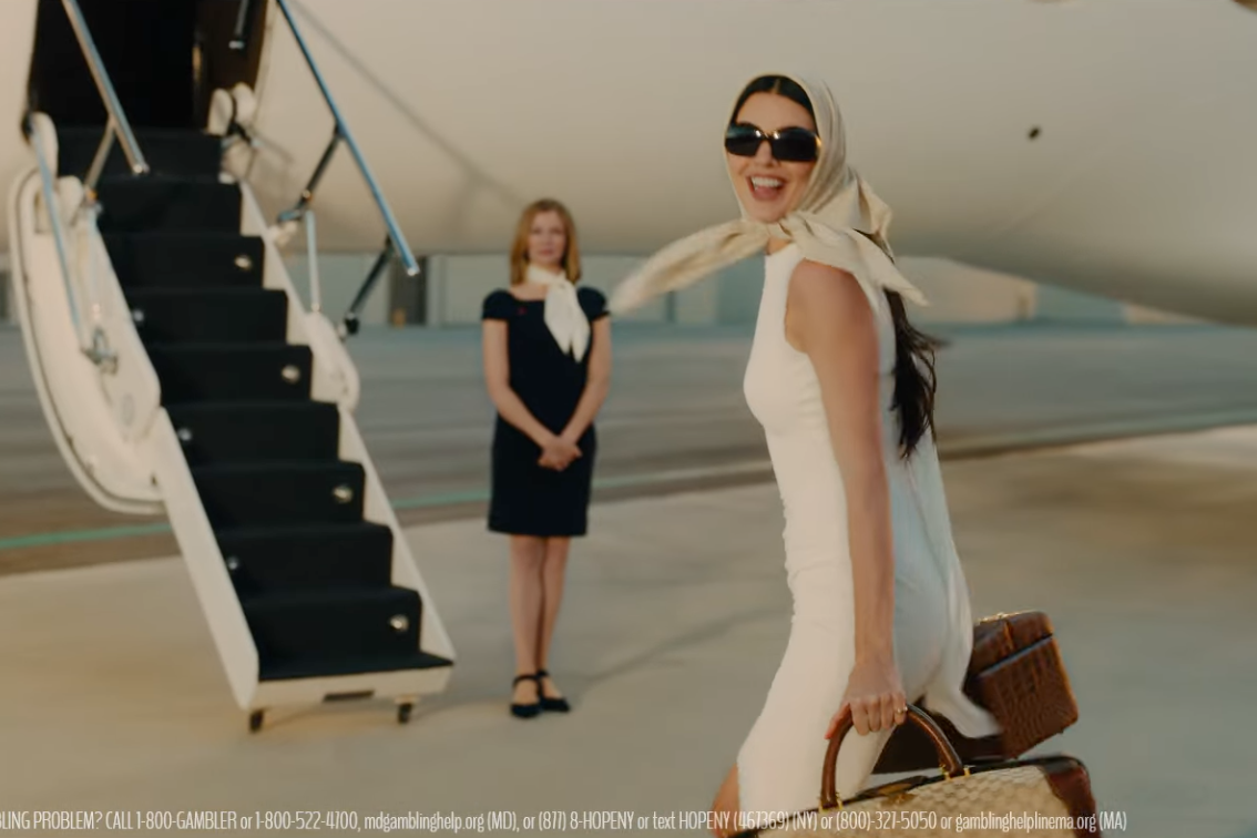 Kendall Jenner in a white dress, sunglasses, and headscarf, holding luggage and smiling, with a private jet and another woman in the background.