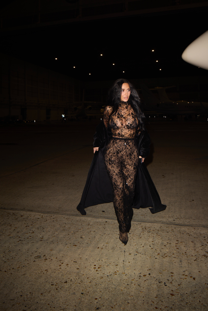 Kim Kardashian in a sheer black lace Balenciaga jumpsuit and a black velvet coat, walking on an asphalt surface at night with a private jet in the background.