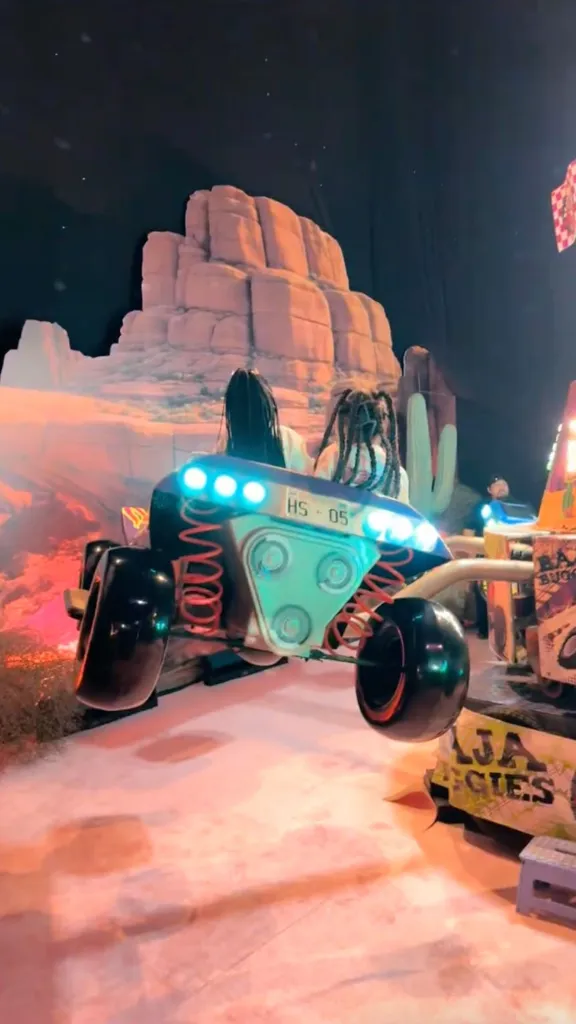 Two people riding in a baja buggy-style amusement park ride with a desert-themed backdrop.