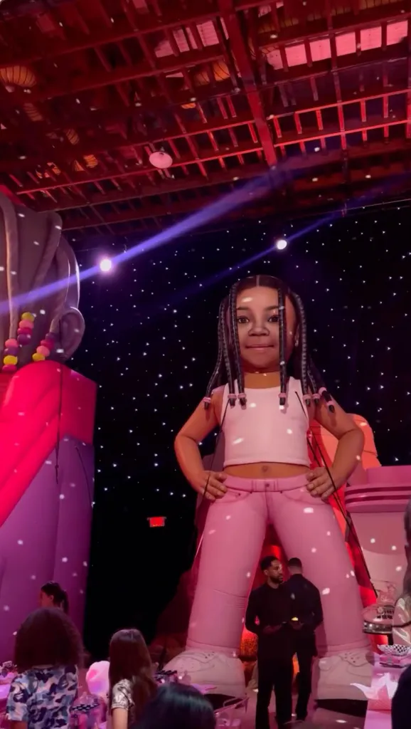 A giant inflatable figure of Stormi with braided hair, wearing a white crop top and pink pants, stands at a birthday party with a starry backdrop.