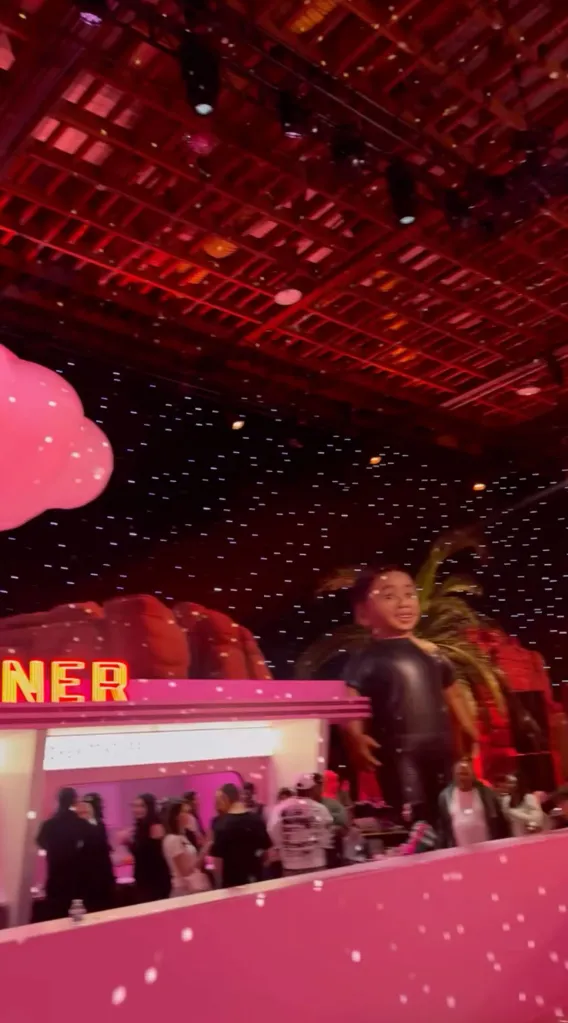 A large indoor birthday party with a pink bar, a giant figure of a child, and a large pink cloud under a black ceiling with white lights.