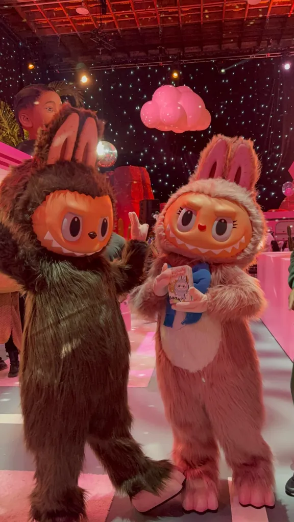 Two individuals in furry monster costumes, one brown and one pink, stand on a pink carpet under a starry ceiling with a pink cloud decoration.
