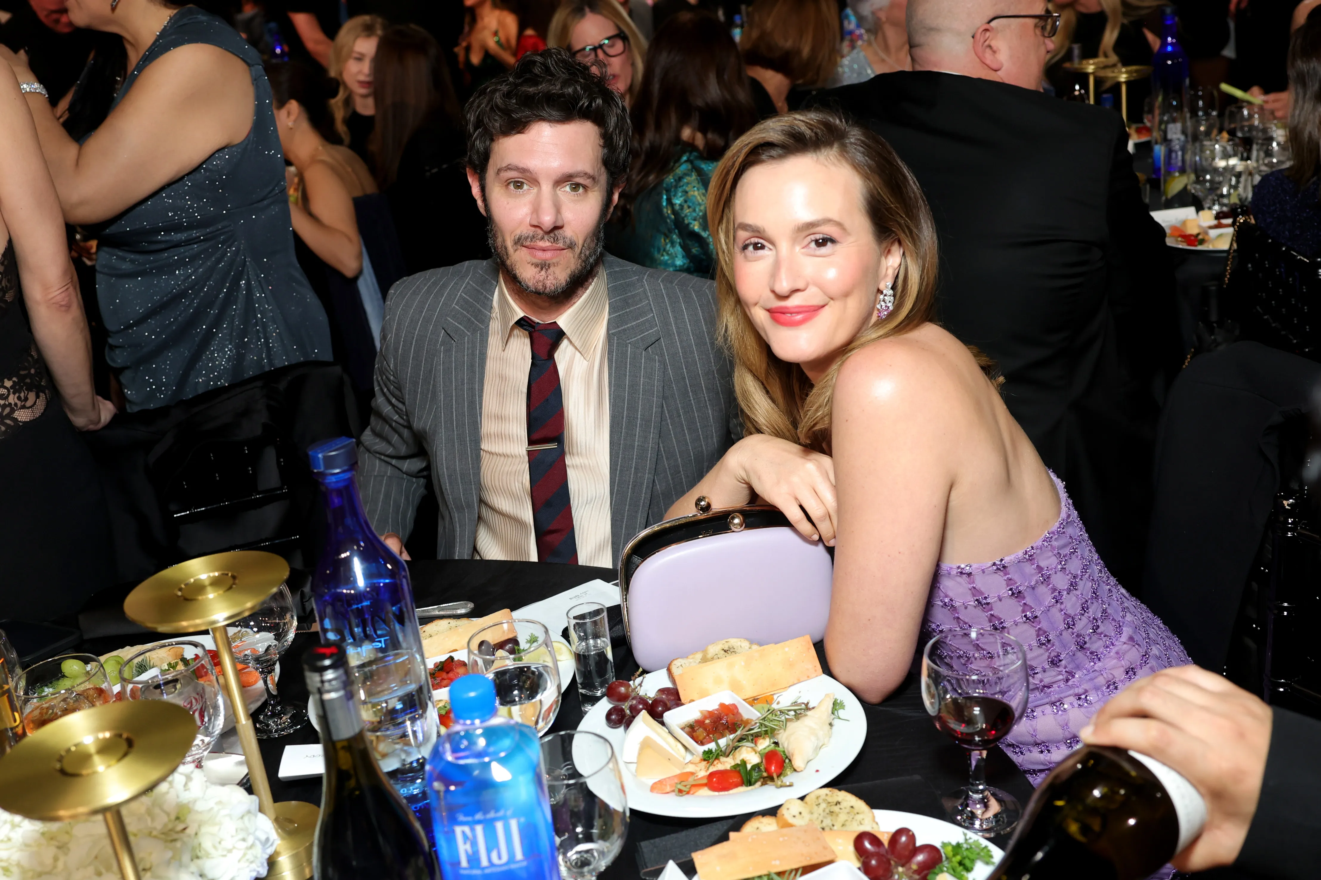Adam Brody and Leighton Meester sitting at a table with plates of food and bottles of Fiji water.