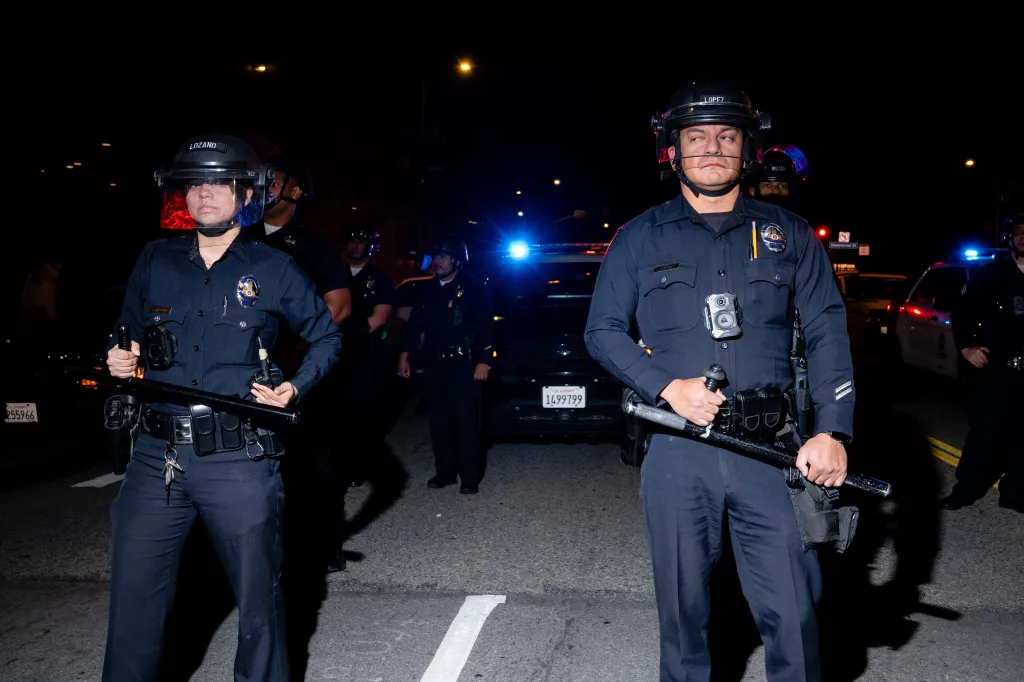 Two police officers in riot gear stand at night with a police car behind them.