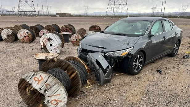 Damaged gray car with a smashed front bumper surrounded by large wooden spools of wire.