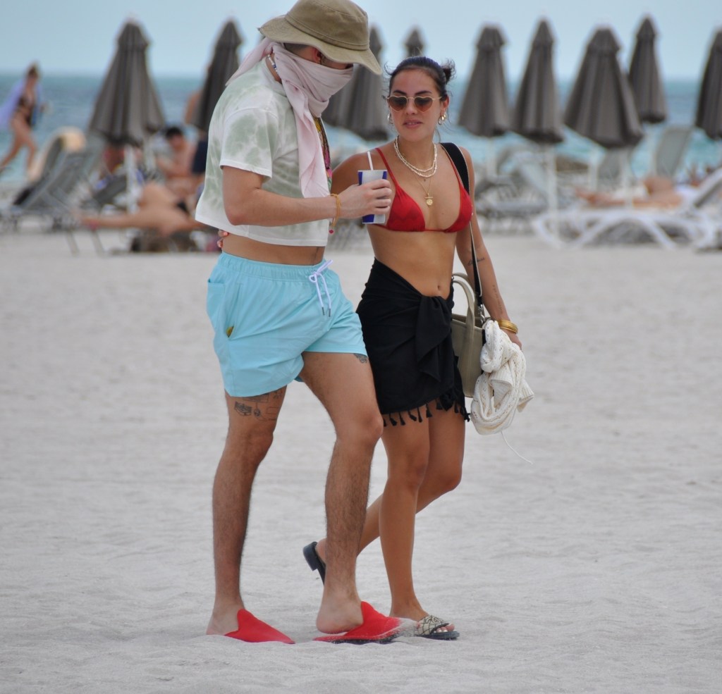Bad Bunny wearing a face covering and Gabriela Berlingeri in a red bikini walking on Miami Beach.