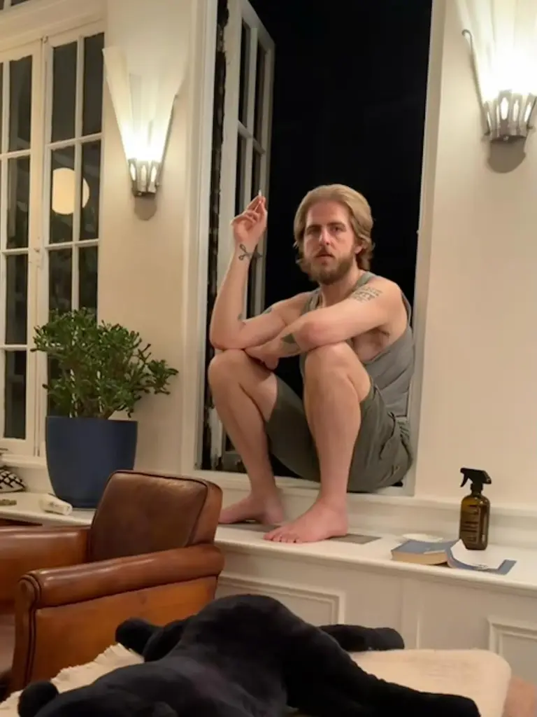 Christian Lee Hutson wearing a gray tank top and green shorts, sits barefoot in a window frame, holding a lit cigarette.