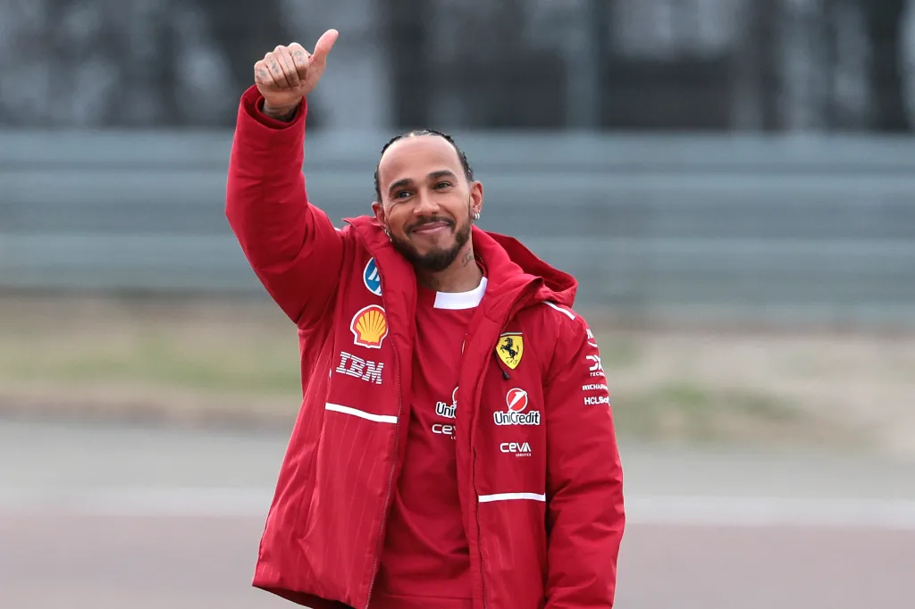 Lewis Hamilton, wearing a red Ferrari jacket, gives a thumbs up.