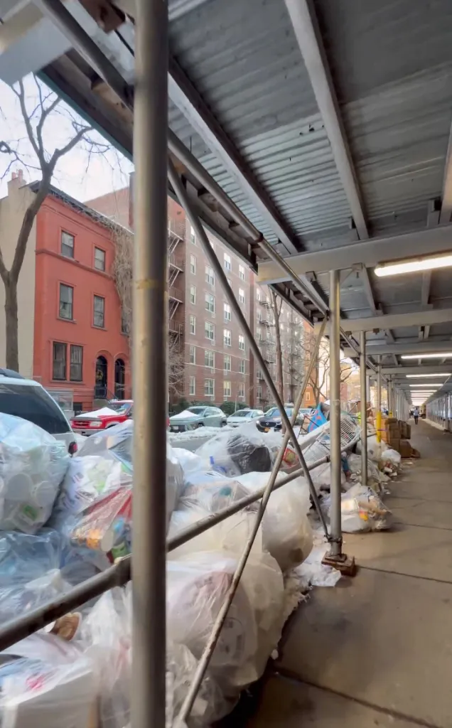 Sidewalk in NYC with piles of bagged garbage under scaffolding.