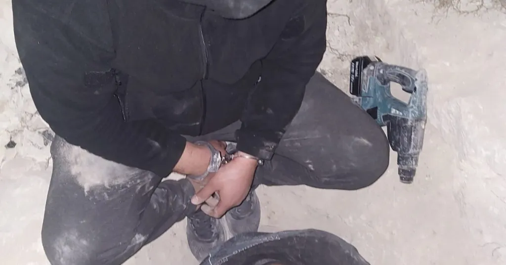A person in handcuffs kneels on dusty ground next to a power tool.