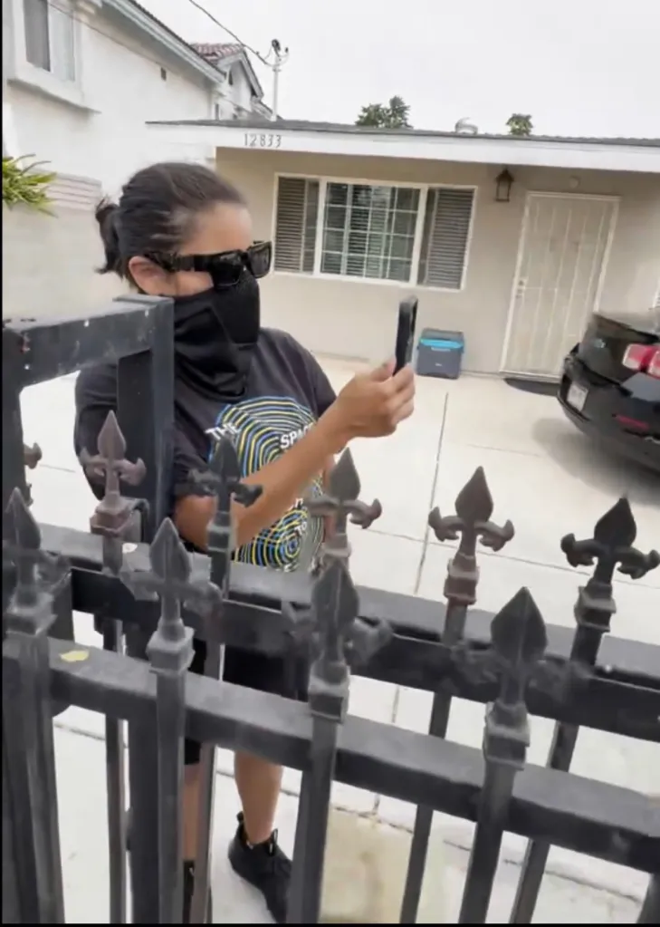 A woman in sunglasses and a black face covering holding a phone to record a house with the number 12833.