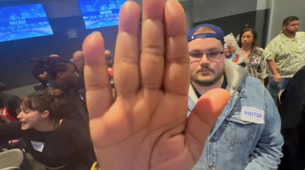 A close-up of a person's outstretched hand at a Los Angeles Police Commission meeting, with multiple people in the background.
