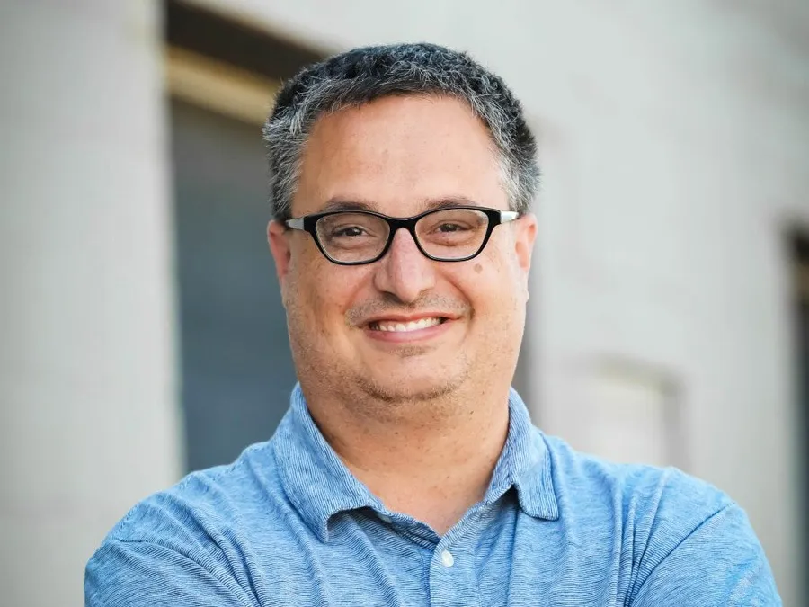 A man with dark, graying hair and black-rimmed glasses smiles at the camera.