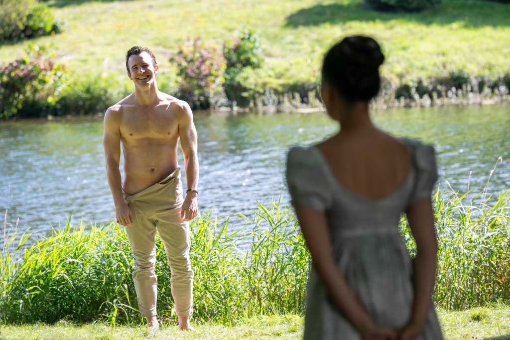 Luke Thompson as Benedict Bridgerton, shirtless with pants around his hips, smiling at Yerin Ha as Sophie Baek, whose back is to the camera, next to a lake.