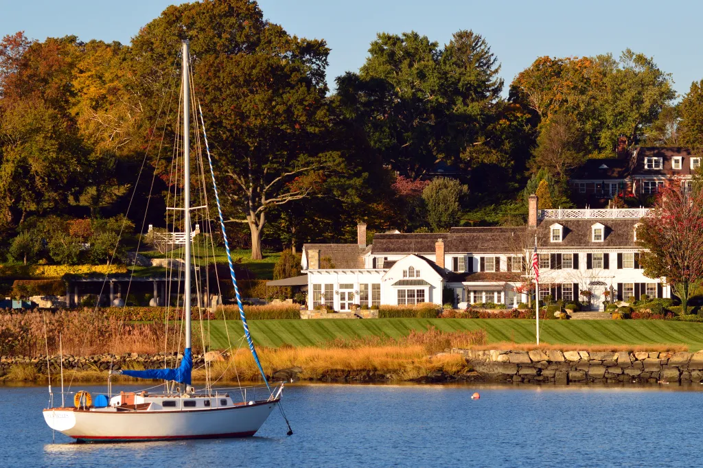 A sailboat on the water in front of luxurious waterfront homes in Greenwich, Connecticut.
