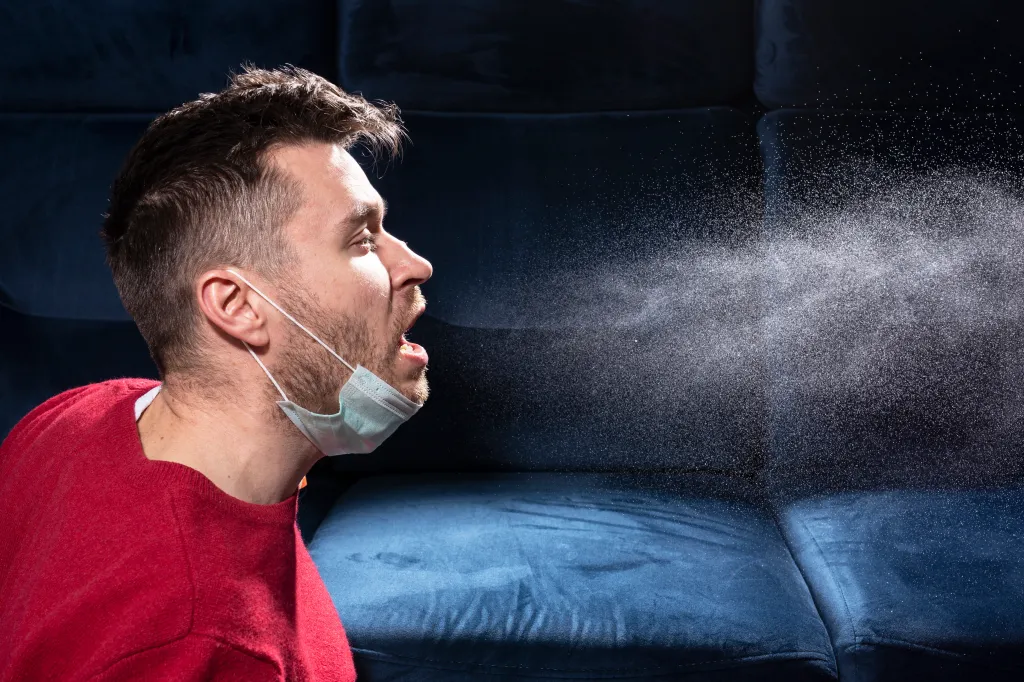 Man with a medical mask pulled down, coughing and releasing a spray of droplets into the air.