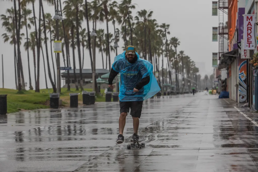A man wearing a blue poncho and baseball cap rides a skateboard on a wet boardwalk, with palm trees and buildings in the background during heavy rainfall.