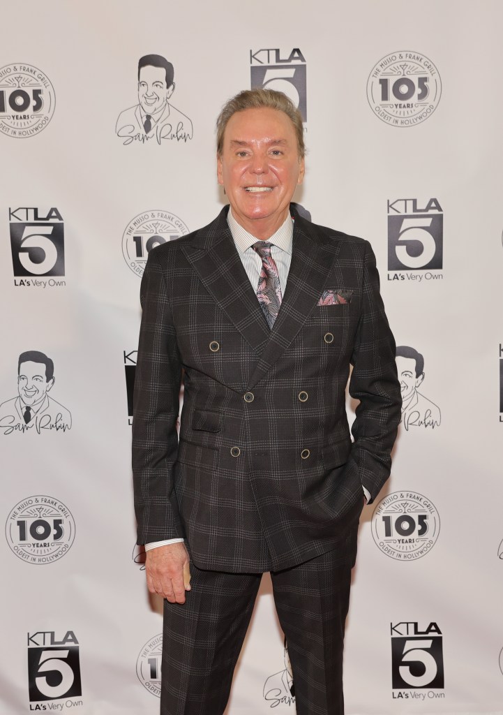 Mark Kriski in a plaid suit and patterned tie, smiling against a white backdrop with event logos.