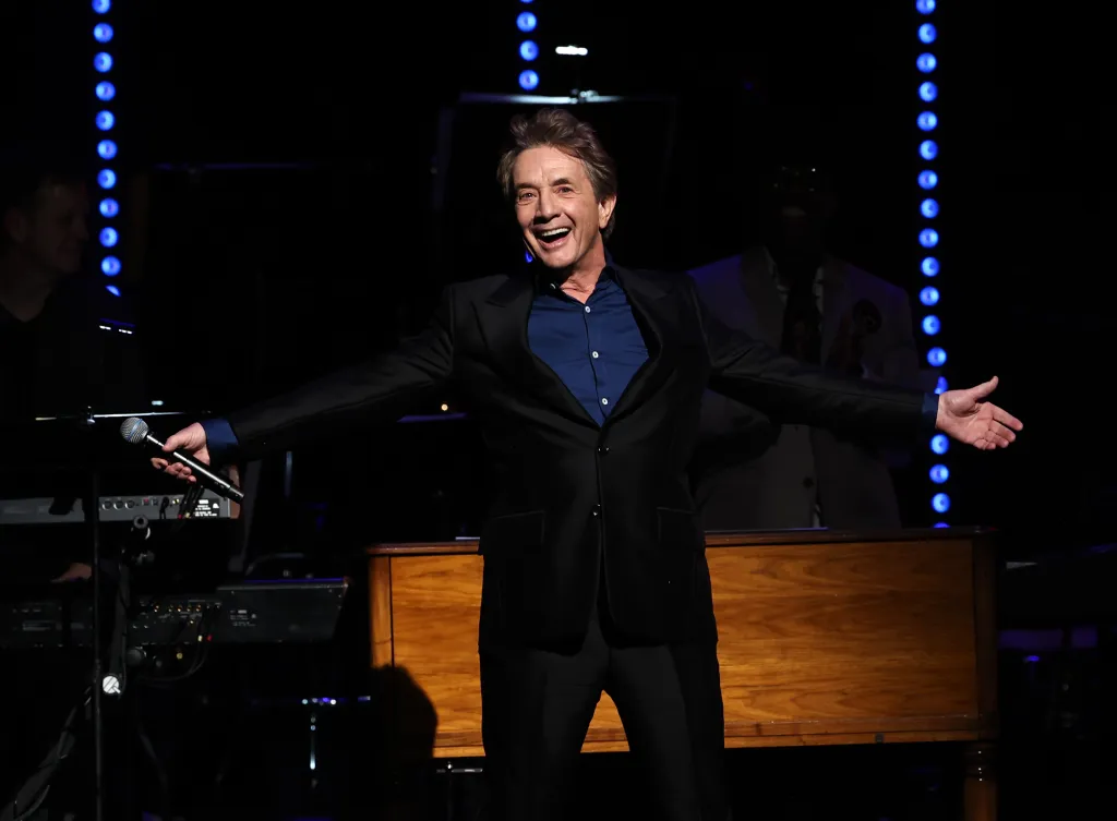 Martin Short onstage speaking with a microphone, arms outstretched, wearing a black suit and blue shirt.