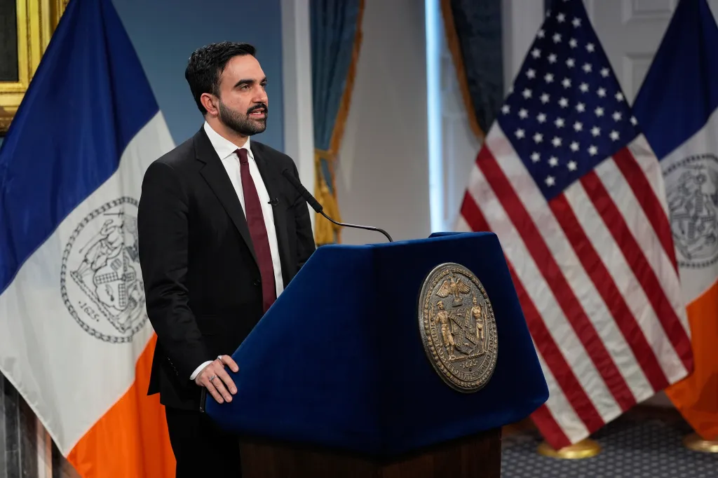 Zohran Mamdani speaks at a news conference behind a blue podium with a seal, flanked by the New York City flag and the American flag.