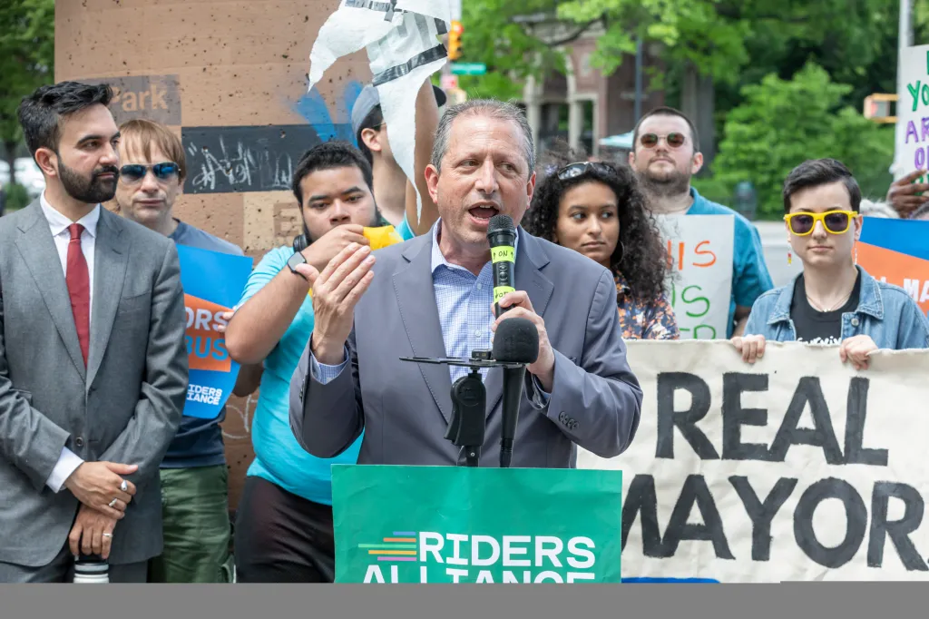 Brad Lander speaking into a microphone at a Riders Alliance rally with Zohran Mamdani and other attendees behind him.