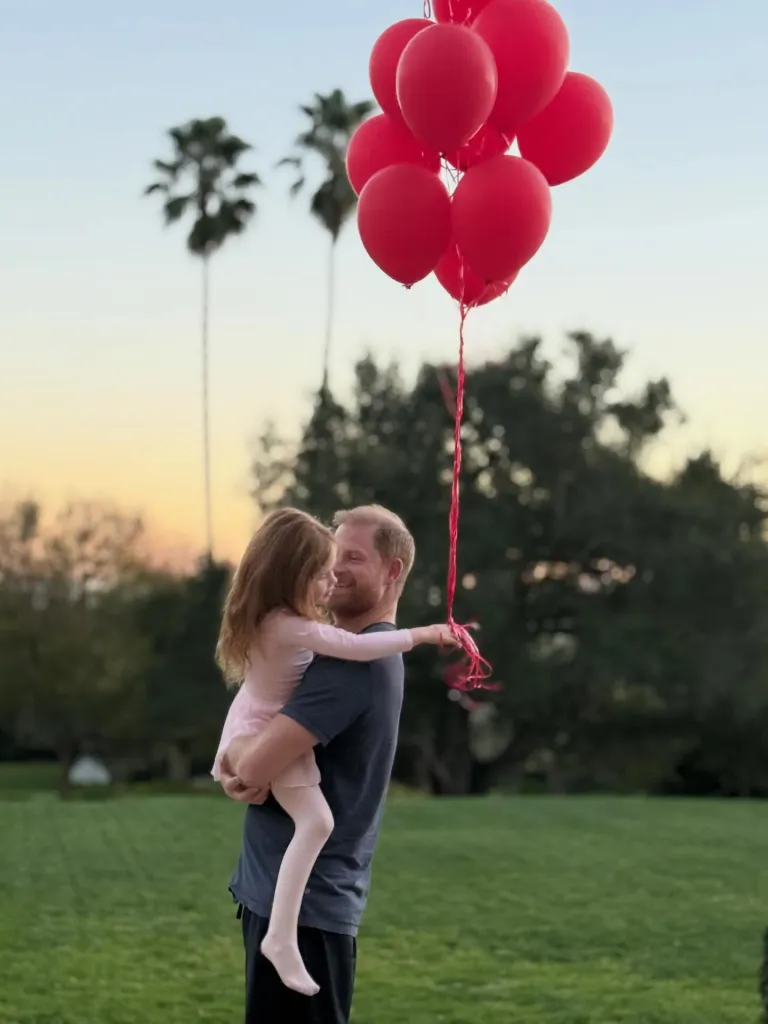 Prince Harry holding a young girl who is holding a bouquet of red balloons.