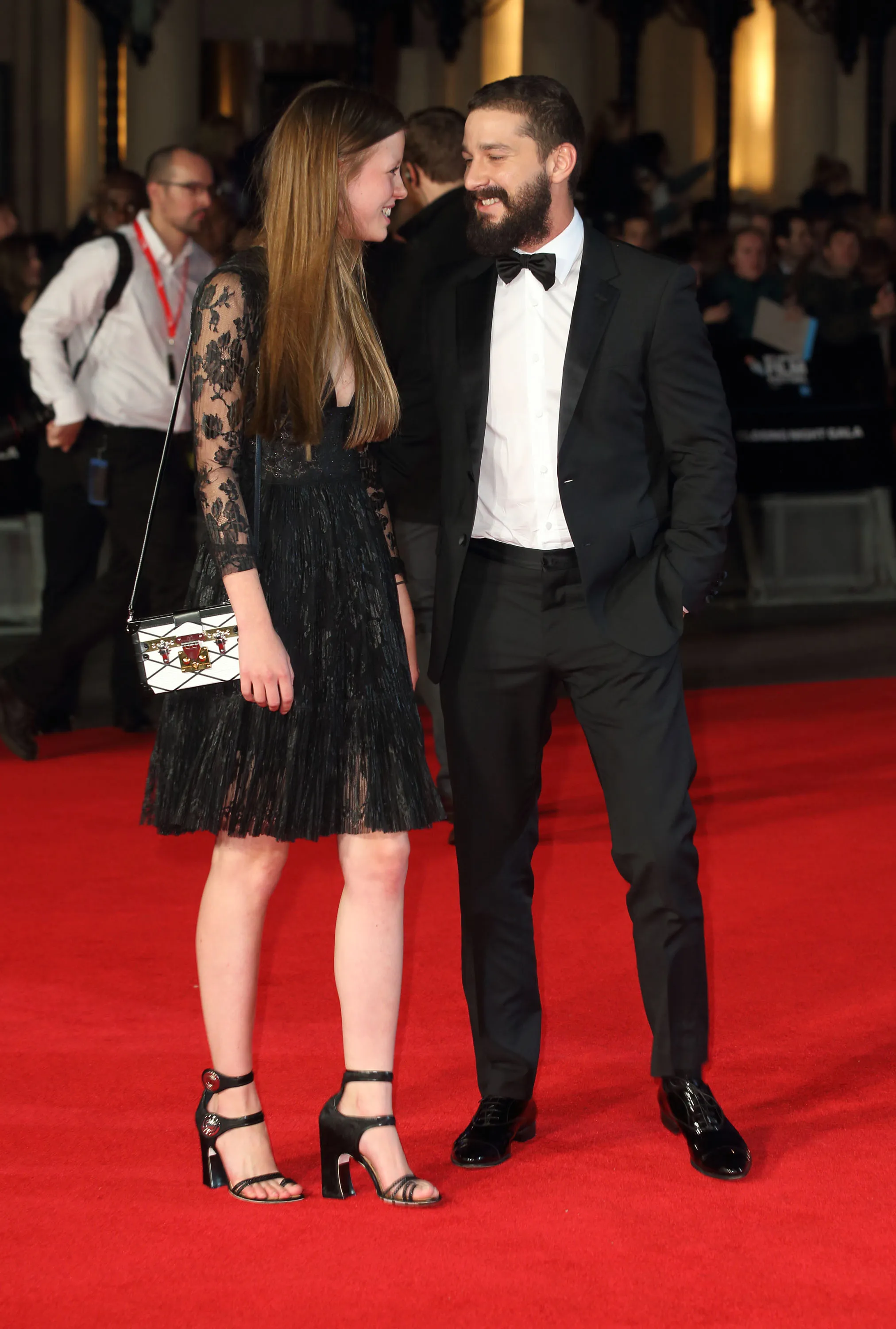Mia Goth and Shia LaBeouf smiling at each other on a red carpet.
