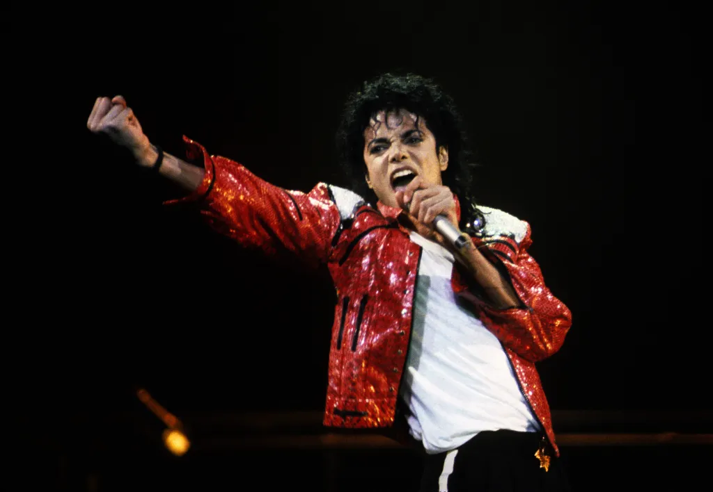 Michael Jackson performing in concert, wearing a red sequined jacket, white t-shirt, and black pants, with one arm raised and holding a microphone.