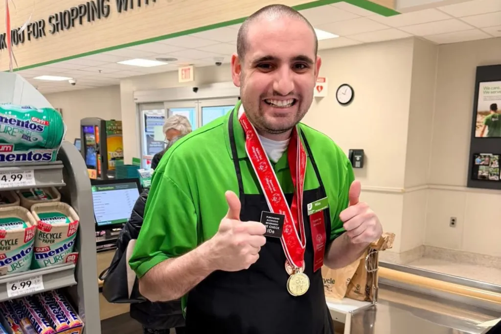Michael Masterangelo at Publix, wearing a green shirt, black apron, and a Special Olympics medal, giving a thumbs up.
