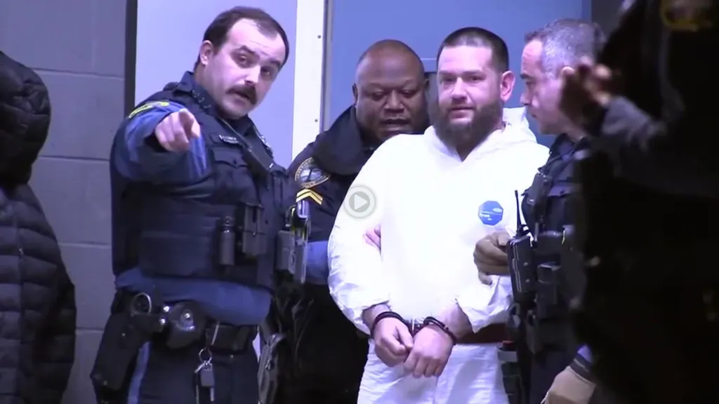 Michael Phillips, handcuffed and wearing a white jumpsuit, being led by police officers.