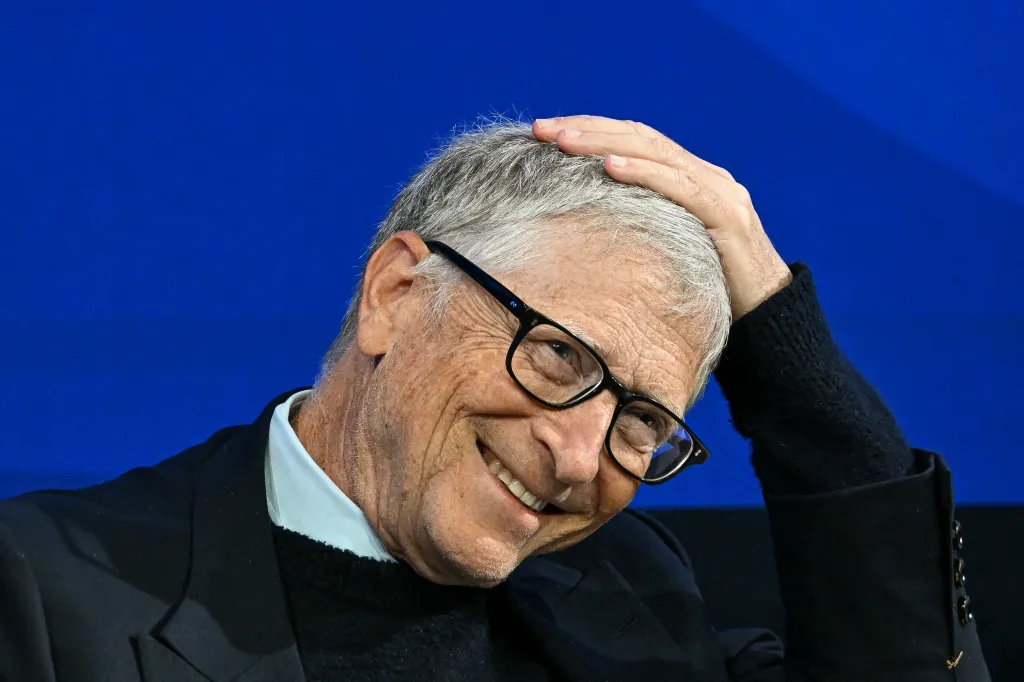 Bill Gates smiling and touching his gray hair, wearing glasses and a black sweater over a light blue collared shirt and black jacket.