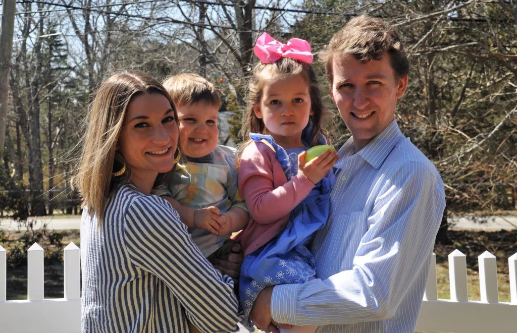 A family of four, including a smiling mother, father, and two young children, stands in front of a bare tree.