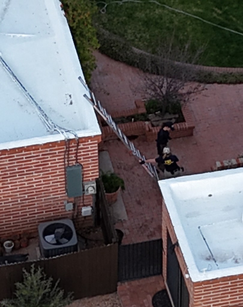 An FBI officer stands on a ladder next to a brick building, while two other officers are on the ground.