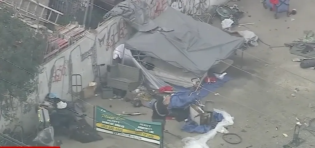 An aerial view of a homeless encampment with tents, tarps, and debris, with a sign in the foreground requesting calls to the Sheriff's Homicide Bureau.