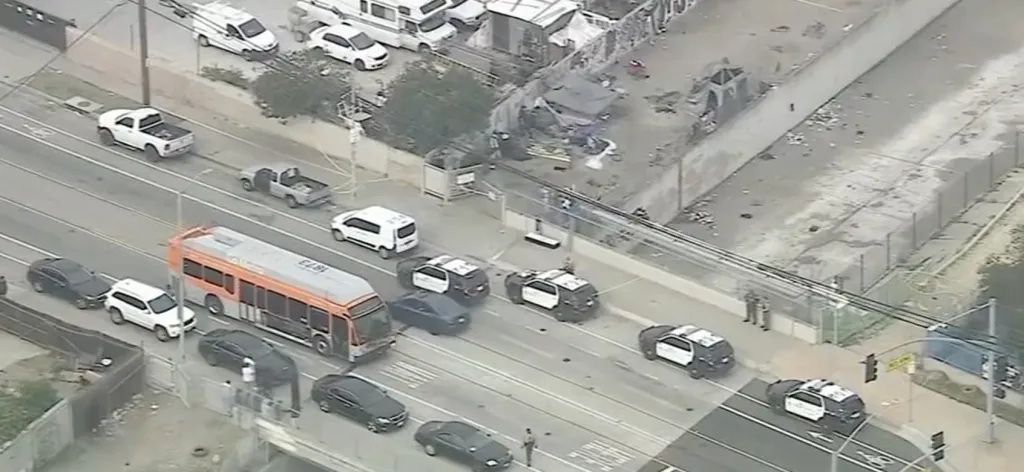 Aerial view of a city street lined with cars and police vehicles, with tents and debris visible on the unpaved shoulder.